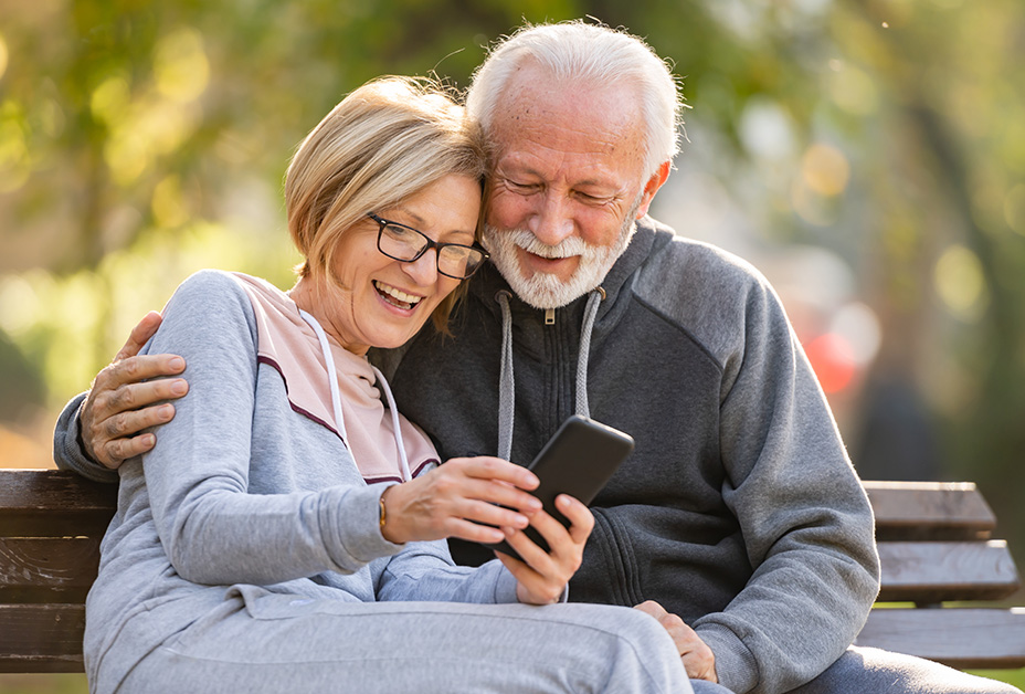 A mature man and women hug and smile while sitting outside on a park bench and looking at a smart phone